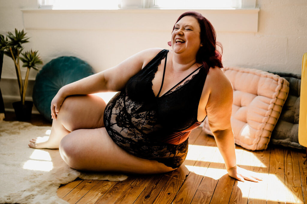 A woman with red hair wearing a black lace bodysuit sits on a wooden floor, smiling and laughing in a sunlit Nashville boudoir filled with soft furnishings and plants in the background.