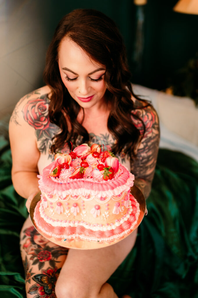 A woman with long brown hair and floral tattoos on her arms and chest holds a pink decorated cake topped with strawberries and cherries, smiling softly in this Nashville boudoir-inspired moment.