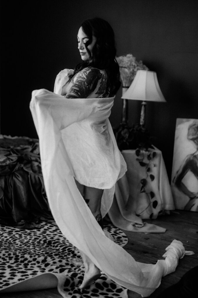A woman with tattoos stands barefoot on a patterned rug, draped in sheer white fabric, posing gracefully in a softly lit Nashville boudoir with dark walls, a bed, a lamp, and artwork. Black and white photo.
