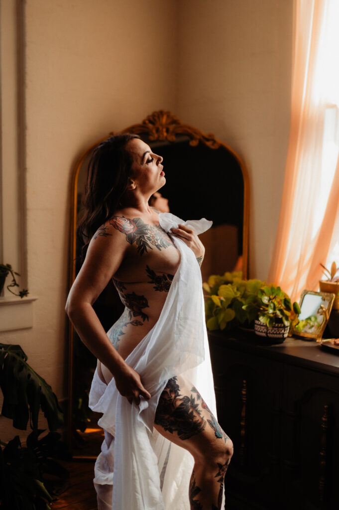 A tattooed woman draped in a white sheet poses by a window, basking in warm sunlight. In true Nashville boudoir style, she stands before a mirror and lush plants, with soft curtains and natural light illuminating the serene scene.