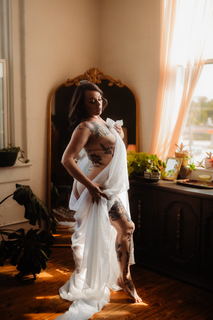 A person with tattoos poses gracefully, draped in a white sheet, standing by a window with sheer curtains. Indoor plants and a large mirror decorate the sunlit room, capturing the elegant charm of a Nashville boudoir.