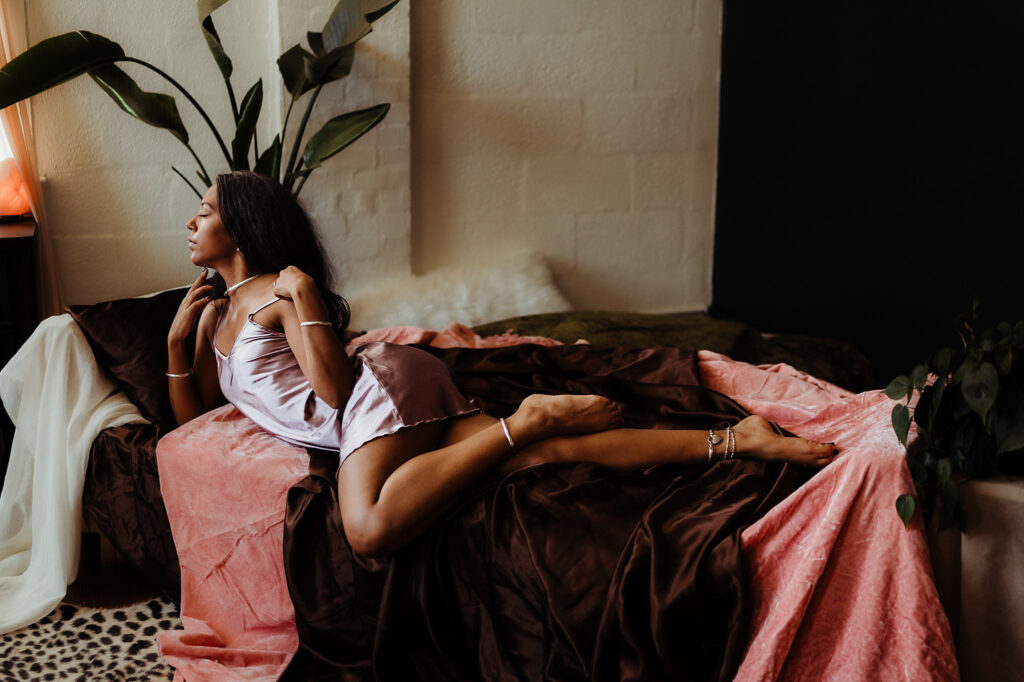 A woman in a satin slip dress reclines on a bed draped with pink and brown fabric, gazing to the side. Soft natural light from the left and lush green plants enhance this elegant Nashville boudoir scene.