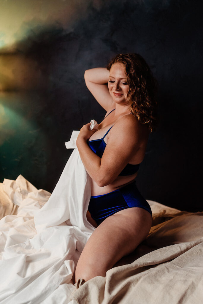A woman with curly red hair sits on a bed wearing blue lingerie, smiling with her eyes closed and holding a white sheet against her chest. The softly lit scene captures the intimate charm of a Nashville boudoir with dark walls in the background.