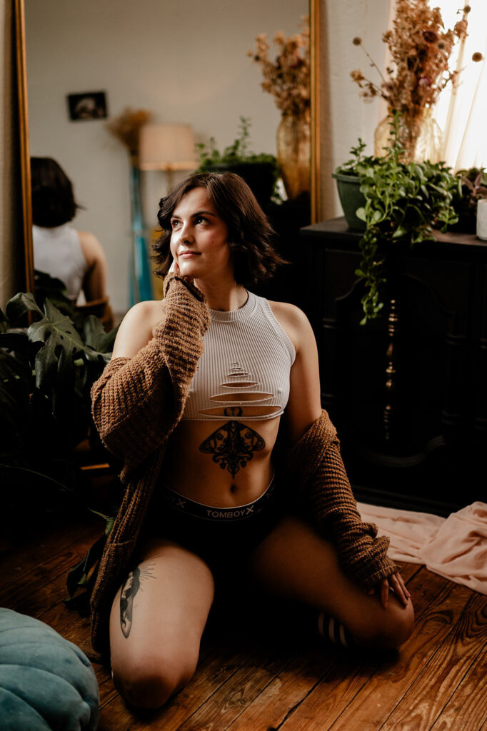 A person with short dark hair kneels on a wooden floor in front of a mirror, wearing a white crop top, black shorts, and a brown cardigan. Plants and dried flowers decorate the cozy, softly lit nashville boudoir.