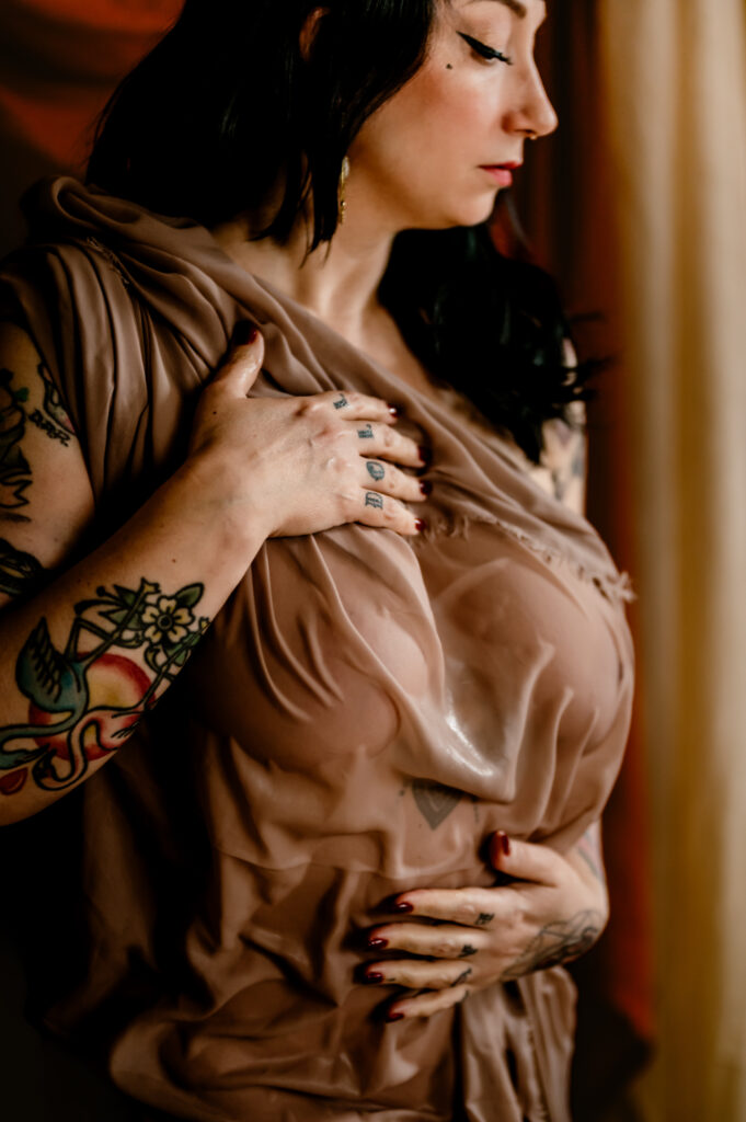 A woman with black hair and tattoos on her arm stands near a window, wearing a sheer, beige fabric draped over her body. In this Nashville boudoir scene, she holds the fabric near her chest and stomach, looking down softly.
