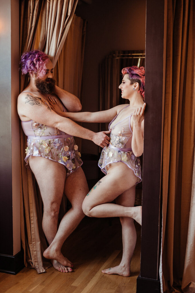 Two people with pink and purple hair stand in front of tan curtains, wearing matching sheer, floral lingerie outfits. Their bodies interact playfully, capturing the intimate vibe of a Nashville boudoir session.