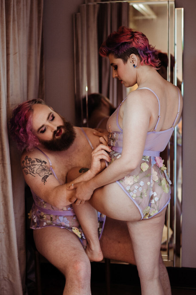 Two people with pink hair, wearing matching sheer lilac lingerie with floral designs, share an intimate, tender moment in front of a mirror—a perfect reflection of connection and beauty, captured in signature Nashville boudoir style.