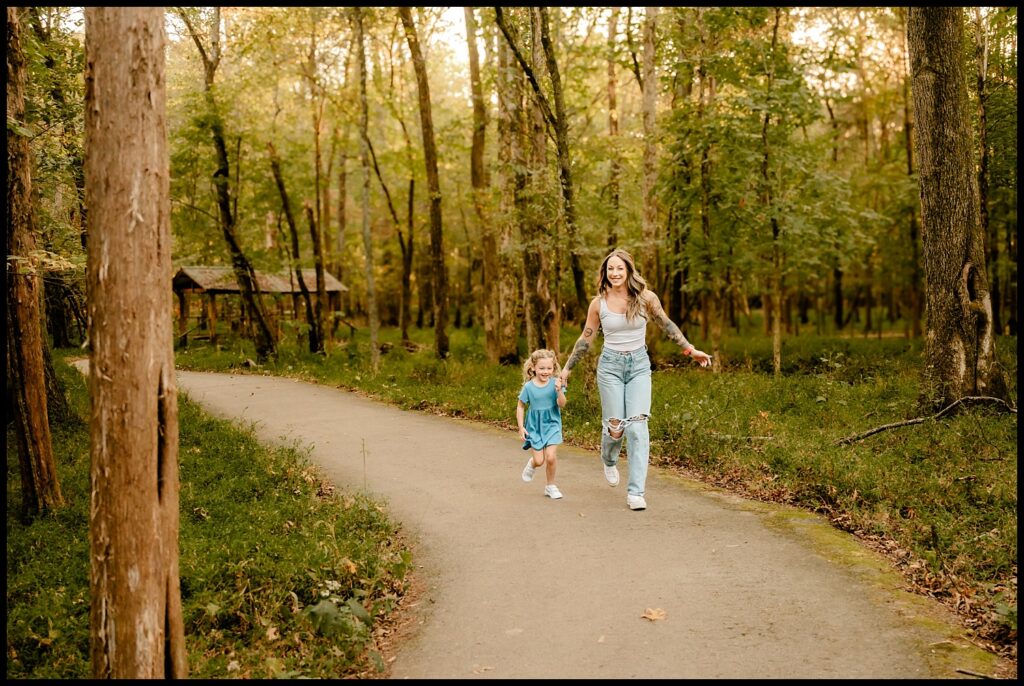 A woman and a young girl holding hands and running joyfully along a sunlit forest path, both smiling brightly. This heartwarming moment is perfectly captured by a Nashville Family Photographer during their Together Session.