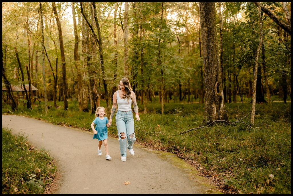 A woman and a young girl walk together on a paved path through a sunlit forest, both smiling and dressed casually. The lush, green setting is perfect for a Nashville Family Photographer to capture one of their joyful Together Sessions.