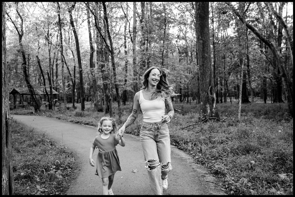A woman and a young girl hold hands and run along a paved path in a forest. Both are smiling and look happy. This black and white moment captures the joy of Family Photography with a Nashville Family Photographer.