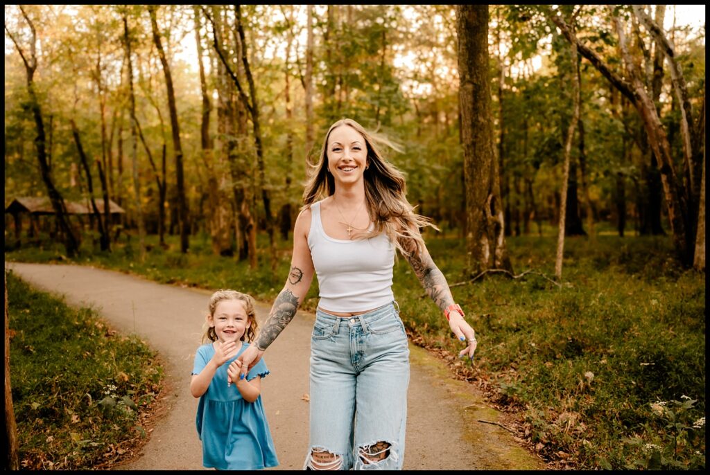 A smiling woman in a white tank top and ripped jeans holds hands with a young girl in a blue dress as they walk together on a sunlit forest path—capturing the joy and connection of a Nashville Family Photographer’s Together Sessions.
