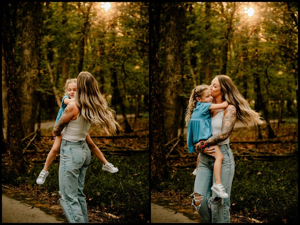 A woman with long hair and tattoos holds and kisses a young girl in a blue dress as they stand outside on a wooded path at sunset—capturing the happy, affectionate bond celebrated by a Nashville Family Photographer.