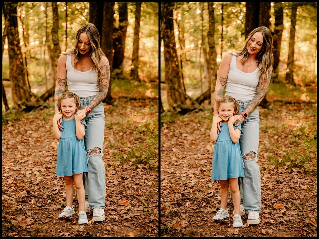 A woman with tattoos, white tank top, and ripped jeans stands in a sunlit forest, smiling and holding a young girl in a blue dress who is grinning; they pose together on a leaf-covered ground—captured by a Nashville Family Photographer.