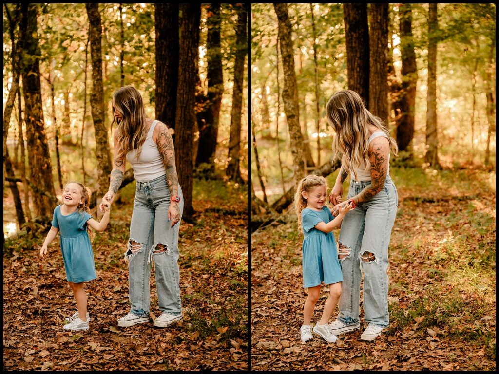 A woman with long hair and tattoos, wearing a white tank top and ripped jeans, laughs and holds hands with a smiling young girl in a blue dress as they stand together in a sunlit forest during a Together Session with a Nashville Family Photographer.
