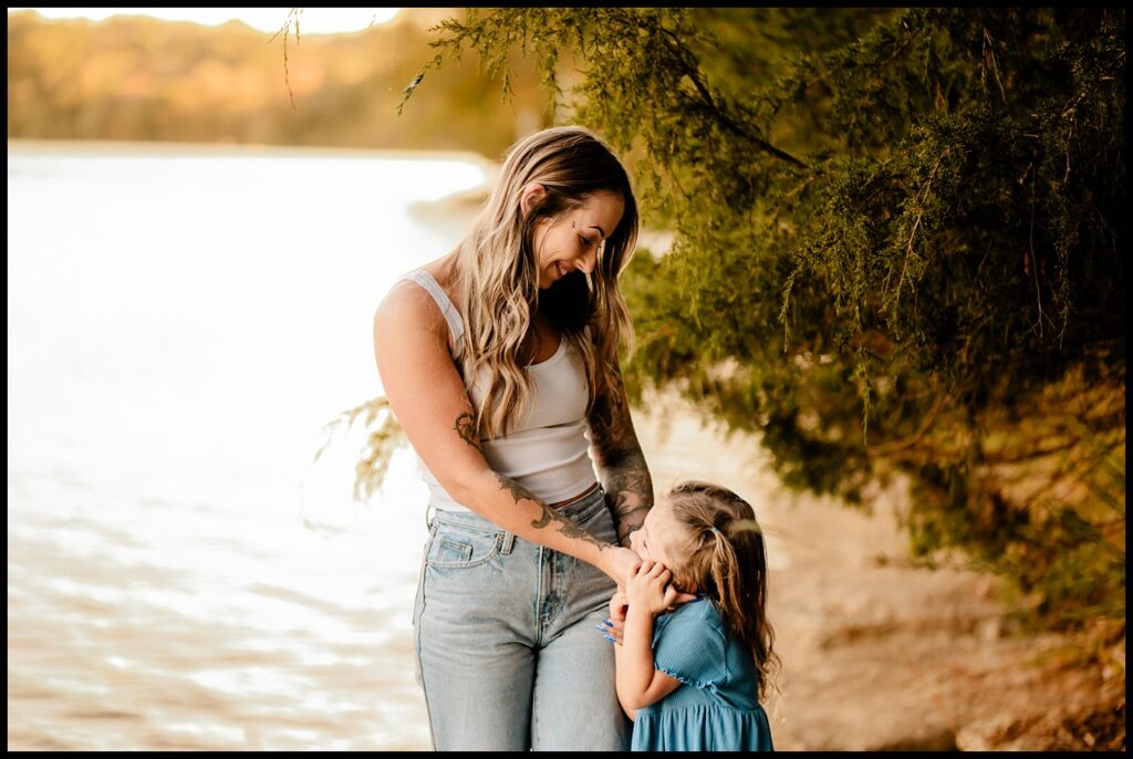 A woman in a white tank top and jeans smiles warmly at a young girl in a blue dress by a lakeside, surrounded by greenery and soft sunlight—capturing a heartfelt moment by a Nashville Family Photographer during Together Sessions.