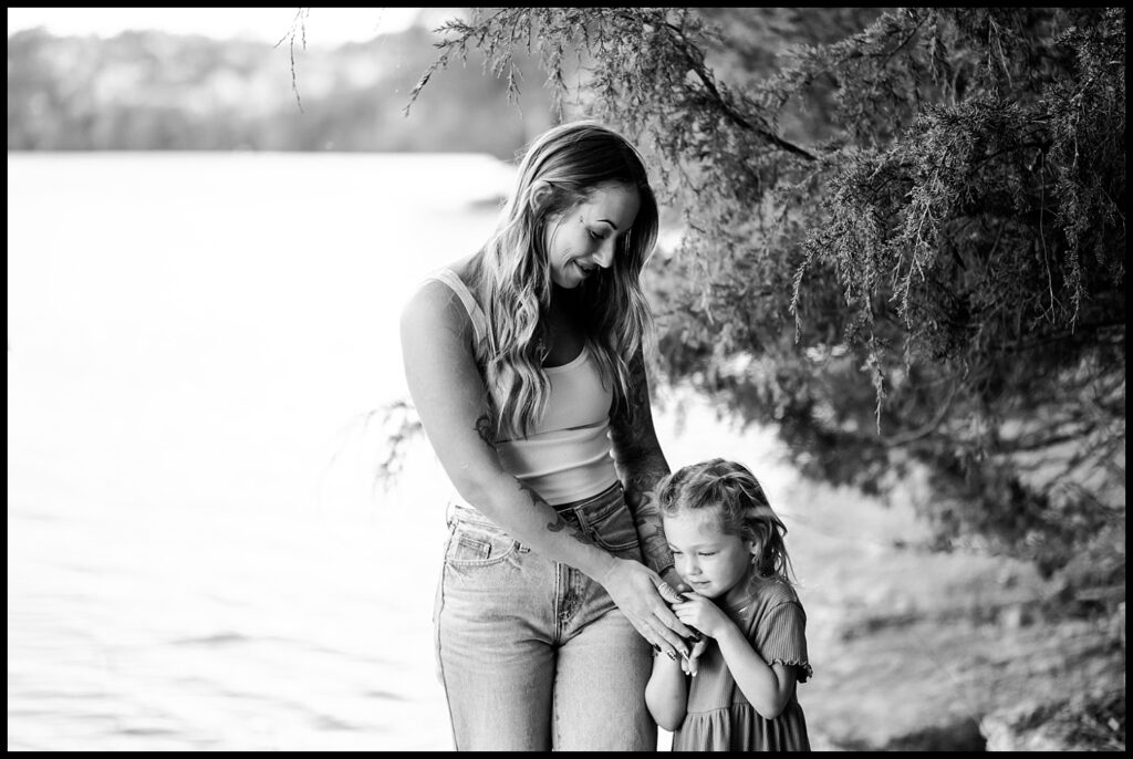 A woman smiles while holding hands with a young girl, who looks down shyly. Outdoors near water and trees, both dressed casually—this black and white moment captures the joy of Together Sessions by a Nashville Family Photographer.