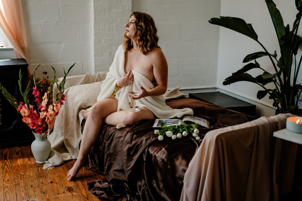 A woman draped in a cream knit blanket sits on a brown satin-covered bed, surrounded by fresh flowers, a lit candle, and an open book, in a softly lit, cozy Nashville boudoir filled with plants and wooden floors.