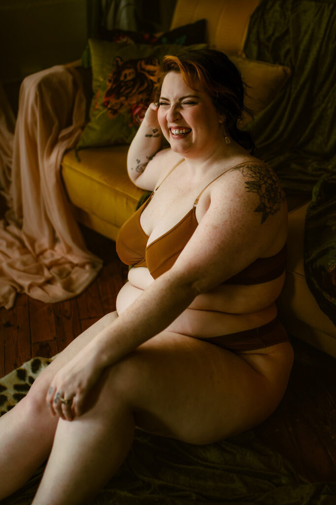 A smiling woman with short hair sits on the floor in brown lingerie, surrounded by draped fabric and a yellow couch with decorative pillows, creating a cozy Nashville boudoir vibe in a warmly lit room.