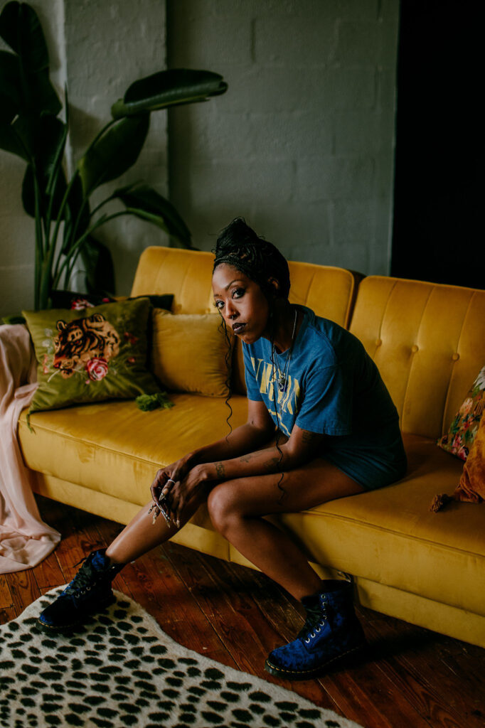A woman in a blue shirt and blue boots sits on a yellow couch with her elbows on her knees, looking at the camera—a chic Nashville boudoir vibe. Decorative pillows and a green plant accent the wooden floor with a spotted rug.