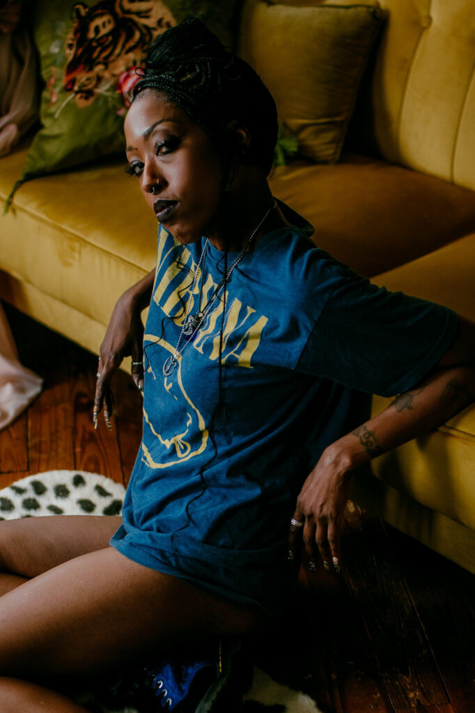 A woman wearing a blue Nirvana t-shirt sits on a rug in front of a yellow couch, looking at the camera with a calm expression. Her hair is styled in braids, she has tattoos on her arms, and the scene captures a relaxed Nashville boudoir vibe.