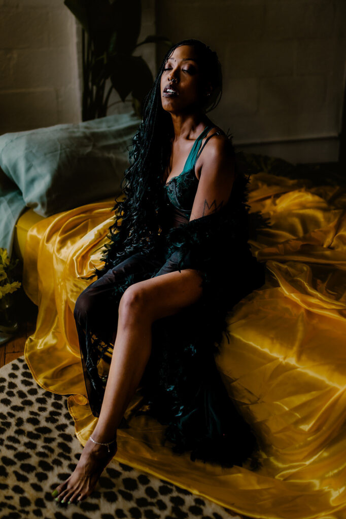 A woman in a dark green dress and black feathered robe sits on a bed with yellow satin sheets, gazing upward. The softly lit room features a plant, a cheetah print rug, and elegant Nashville boudoir vibes.