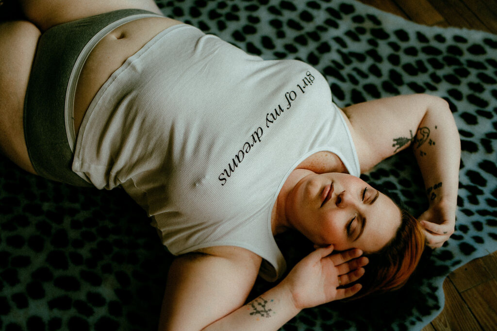 A person with short hair lies on a leopard print blanket, wearing a white tank top with text and gray underwear. Capturing relaxed vibes with eyes closed, this scene channels classic Nashville boudoir style and effortless confidence.