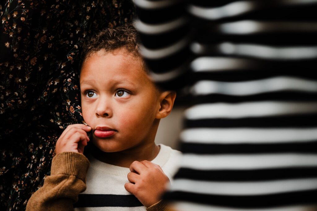 A young boy with curly hair and a concerned expression stands among adults, holding his sweater and touching his face. Captured during a fall family session by a Nashville portrait photographer, he wears a cream and black striped sweater.