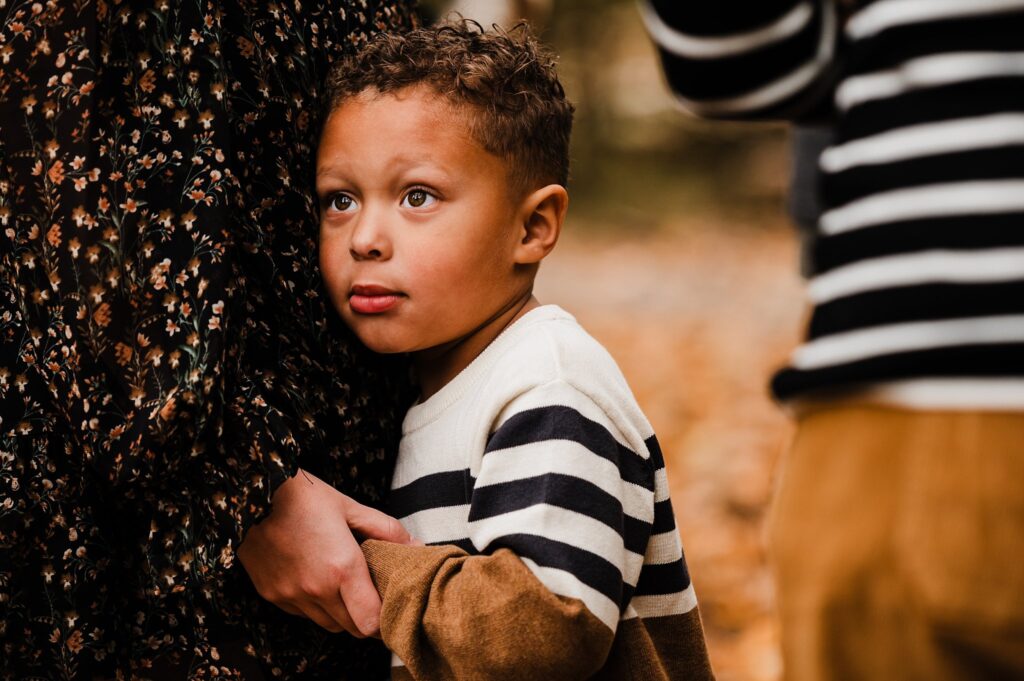 A young boy with curly hair and a striped sweater hugs an adult, looking up with a thoughtful expression. Autumn leaves cover the ground in this cozy fall family session, captured by a Nashville portrait photographer.