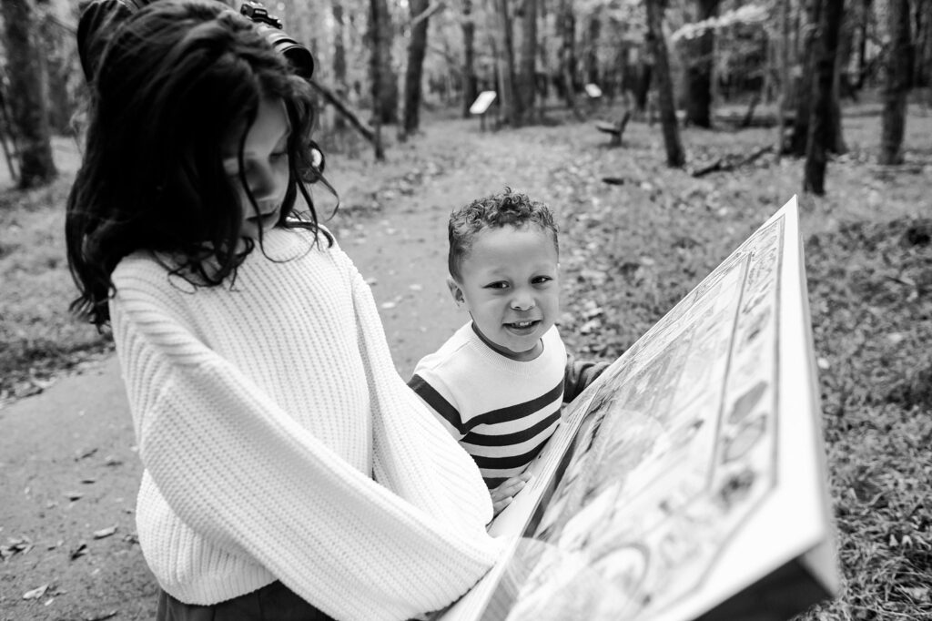 Two children stand on a forest path looking at a large informational sign during a fall family session. One wears a light sweater and looks down, while the other smiles at the camera—capturing a sweet moment in family photography.