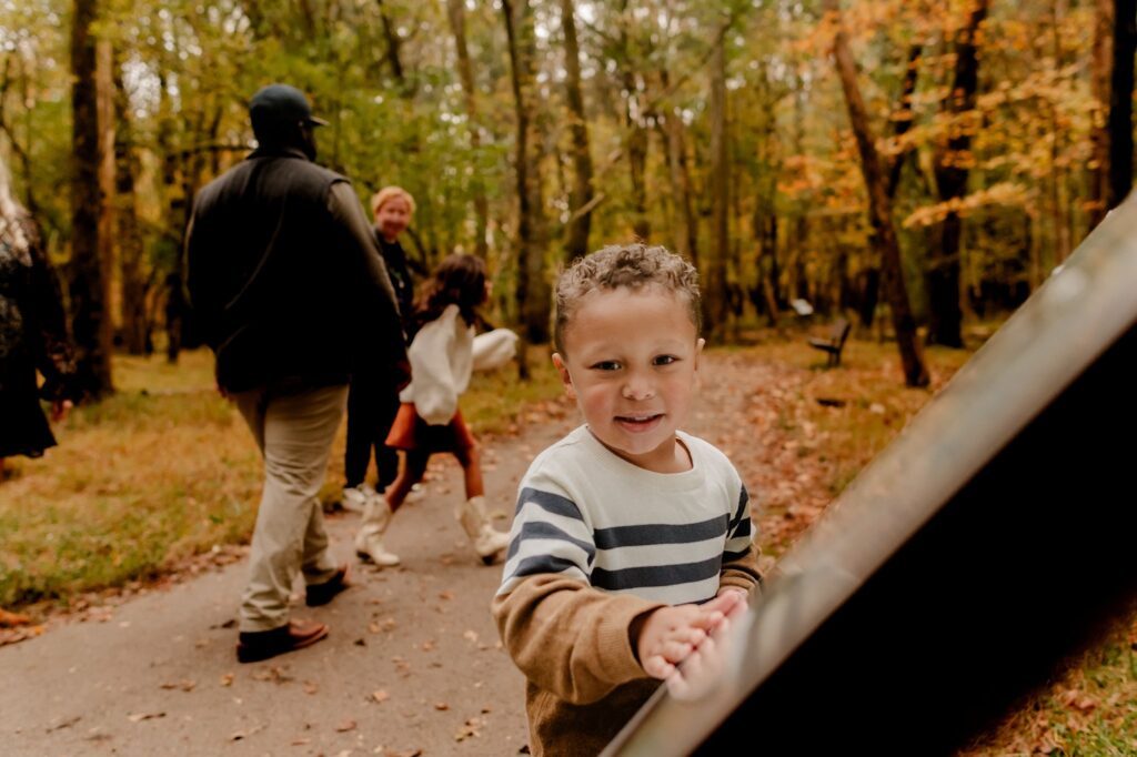 A young boy in a striped sweater smiles at the camera while touching a sign. Behind him, people walk on a forest path surrounded by autumn trees—an ideal scene for a fall family session with your Nashville portrait photographer.