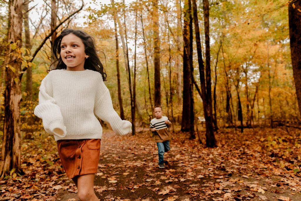Two children run along a leaf-covered path in a forest during autumn, surrounded by colorful trees. Perfect for a fall family session, this joyful moment showcases the warmth and charm captured by a Nashville portrait photographer.