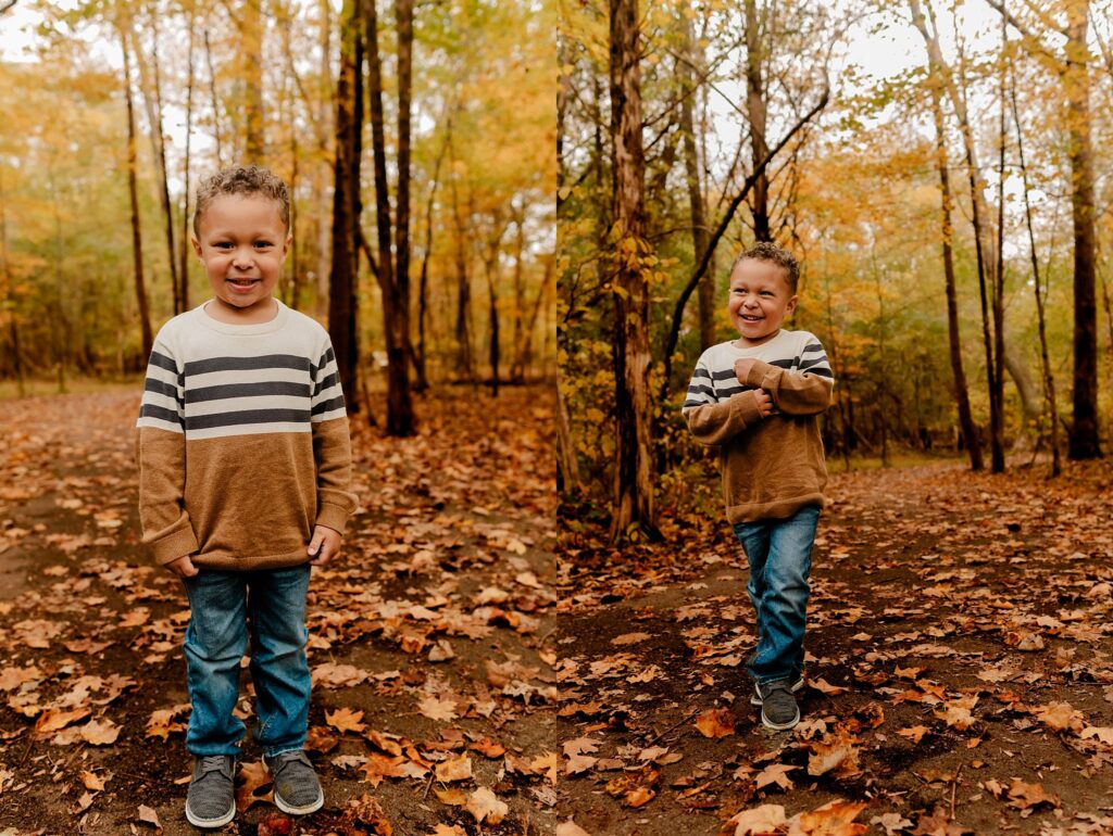 A young boy with curly hair smiles and laughs in an autumn forest during a fall family session, wearing a brown and white striped sweater, blue jeans, and gray shoes. Fallen leaves cover the ground and colorful foliage surrounds him.