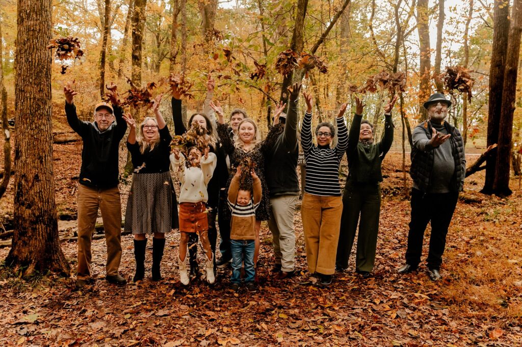 A group of people stand in a forest with autumn leaves, smiling and joyfully tossing leaves into the air. Two children stand in front, the ground covered in orange and brown fallen leaves—perfect for a fall family session.