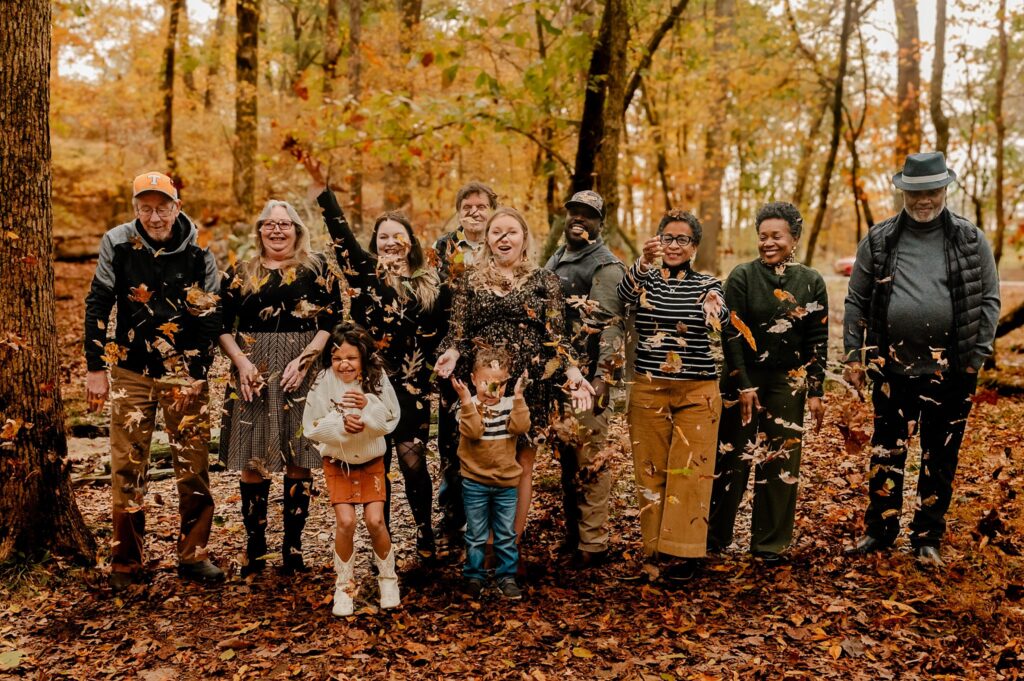 A group of people stand in a forest with autumn leaves on the ground, smiling and throwing leaves into the air during a joyful fall family session captured by a Nashville portrait photographer. The trees display vibrant fall colors.