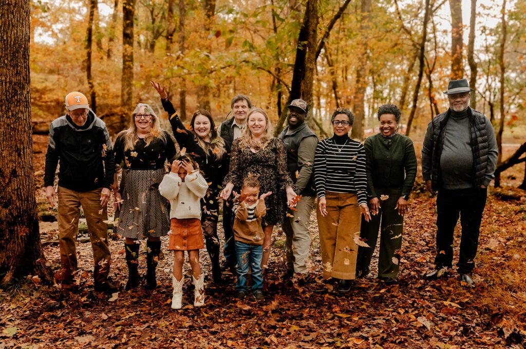 A group of ten people, including children and adults, stand together in a forest with autumn leaves on the ground during a fall family session. Some are smiling and tossing leaves into the air, captured by a Nashville portrait photographer.