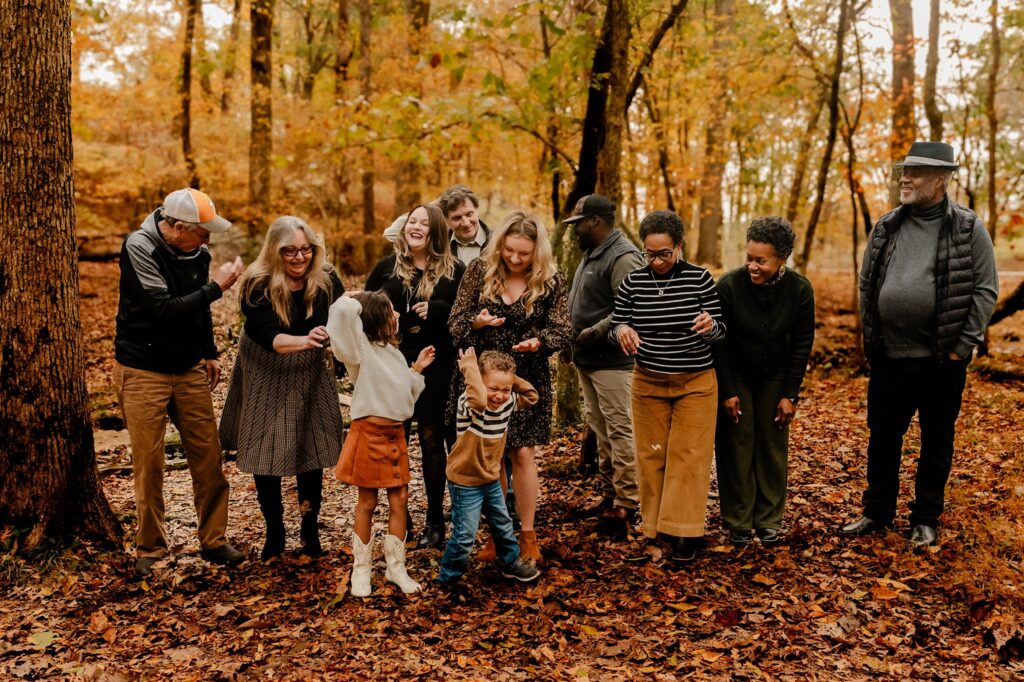 A group of people, including adults and children, stand together outdoors in a forest with autumn leaves on the ground, smiling and laughing while tossing leaves in the air during a fun fall family session.