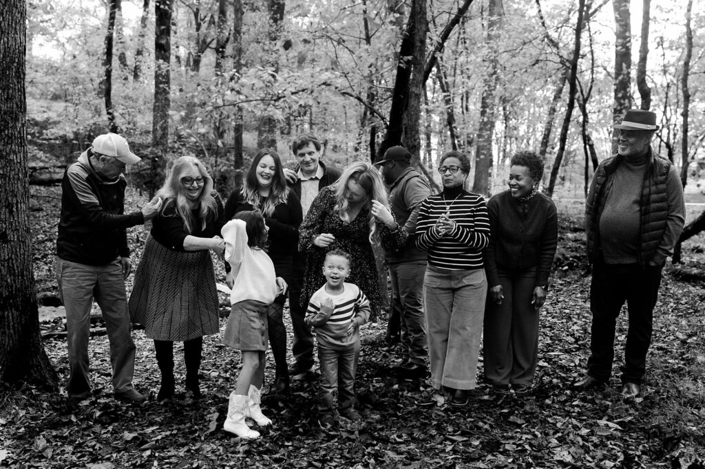 A group of ten people, including adults and two children, stand together in a forest, smiling and laughing on leaf-covered ground during a joyful fall family session—capturing candid moments perfect for family photography.