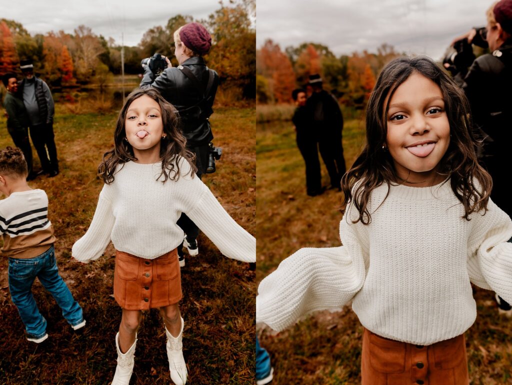 A young girl with long dark hair, wearing a white sweater and brown skirt, playfully sticks out her tongue outdoors during a fall family session. Other people stand in the background on a grassy field with autumn trees.