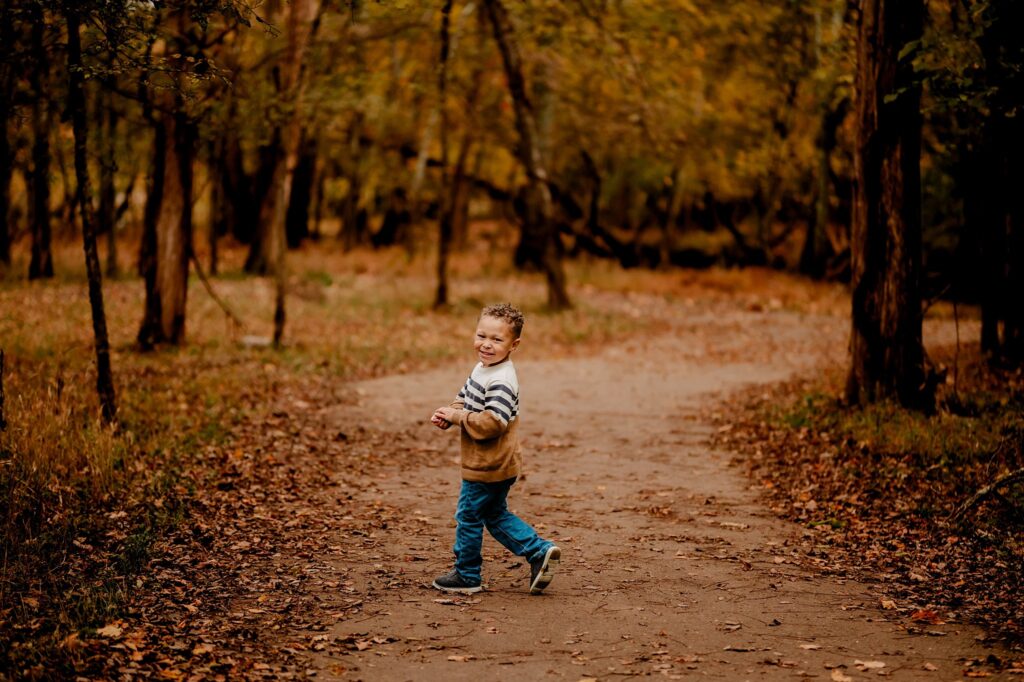 A young boy wearing a striped sweater and blue jeans walks on a leaf-covered path through a forest with autumn foliage, smiling and looking back over his shoulder during a fall family session with a Nashville portrait photographer.