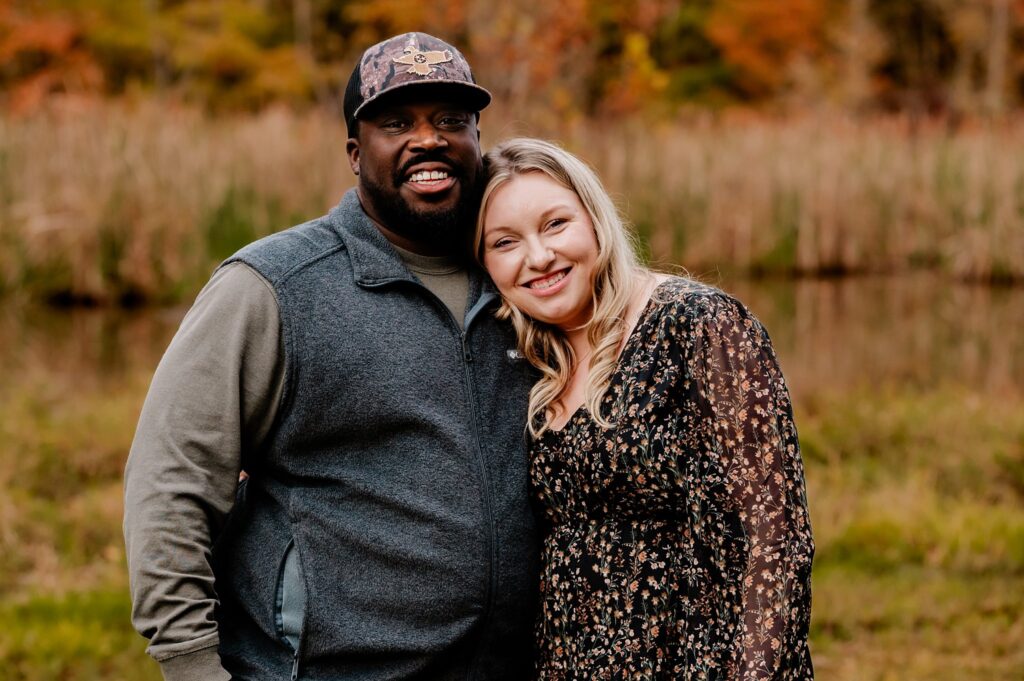 A smiling man and woman stand close together outdoors in front of tall grass and autumn trees, with the woman resting her head on the man’s shoulder during a fall family session by a Nashville portrait photographer.