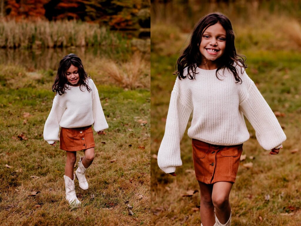 A young girl with long dark hair, wearing a white sweater, brown skirt, and white boots, smiles and walks outside on grass with autumn trees and a pond in the background—perfect for a fall family session with your Nashville portrait photographer.