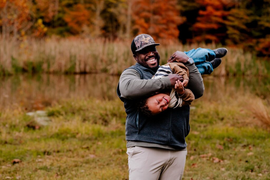 A smiling man in a cap playfully holds a laughing child upside down outdoors, with tall grass and autumn-colored trees setting the scene for a joyful fall family session.