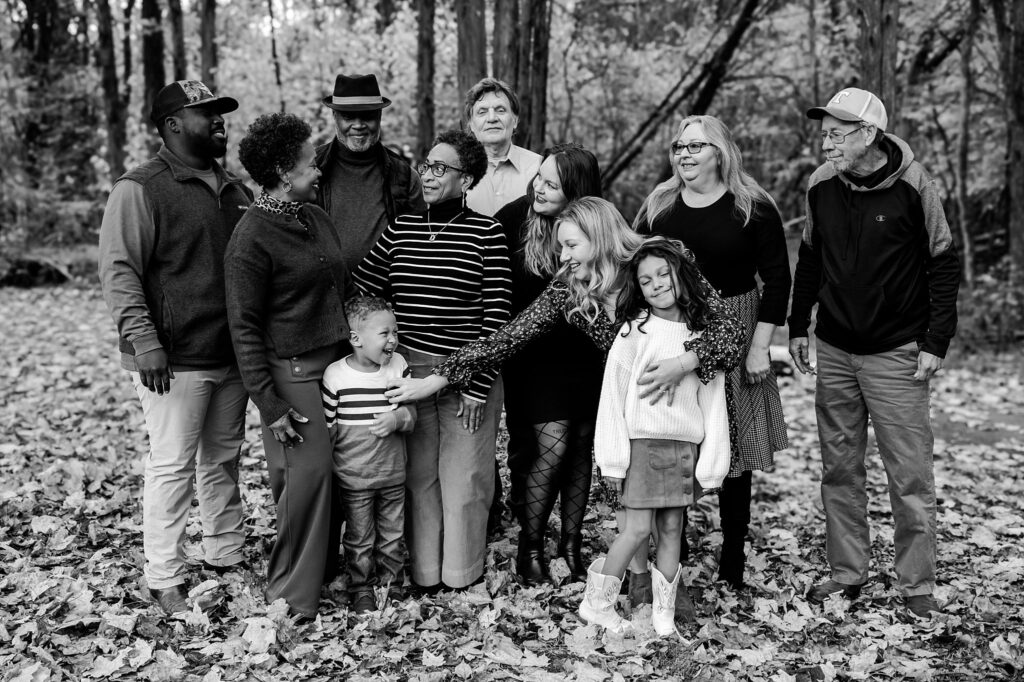A group of eleven people, including adults and children, stand close together outdoors on fallen leaves, smiling and interacting with each other in a forest setting. This black-and-white image captures the warmth of a fall family session by a Nashville portrait photographer.