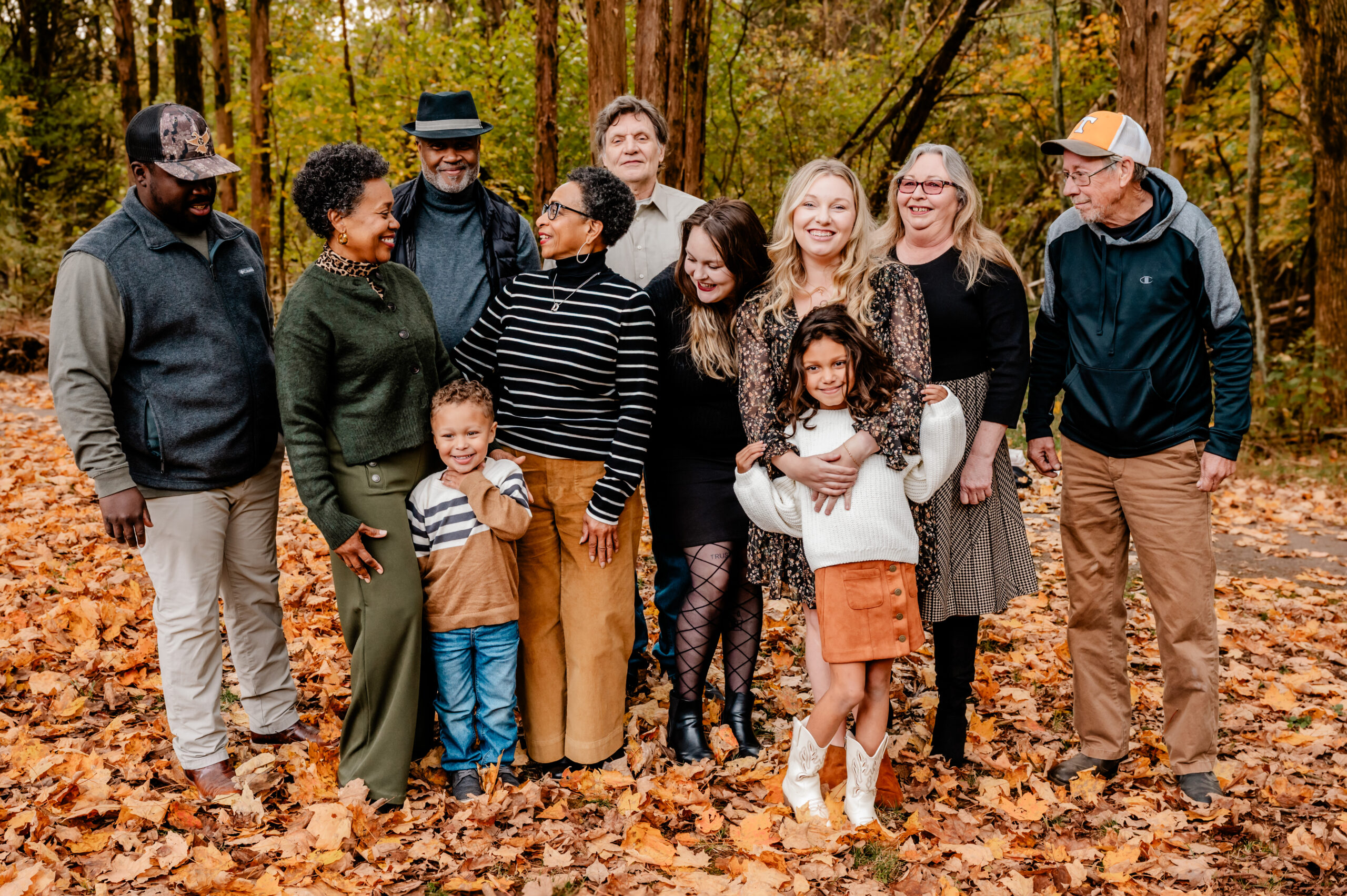 A diverse group of adults and children stand together smiling in a forest with autumn leaves, dressed in warm, casual fall clothing. Captured by a Nashville family photographer, the scene features tall trees and vibrant foliage in the background.