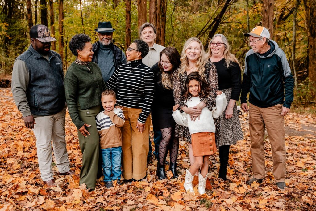 A diverse group of adults and children stand together outdoors on fallen autumn leaves, smiling and laughing, surrounded by tall trees with colorful fall foliage during a vibrant fall family session.