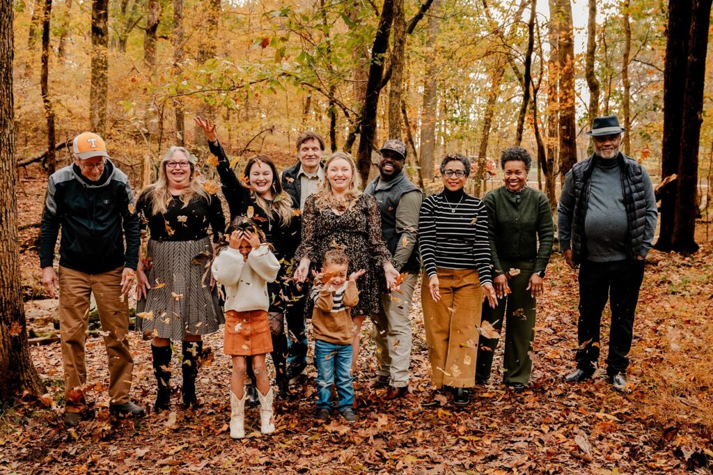 A group of nine adults and two children stand together in a forest with autumn leaves on the ground. Captured by a Nashville portrait photographer, several people are smiling and tossing leaves in the air, surrounded by colorful fall trees.