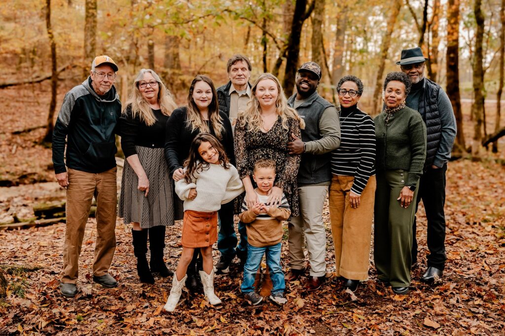 A group of eleven people, including adults and two children, stand close together, smiling in a forest with autumn leaves on the ground—perfect for a fall family session captured by a Nashville portrait photographer.