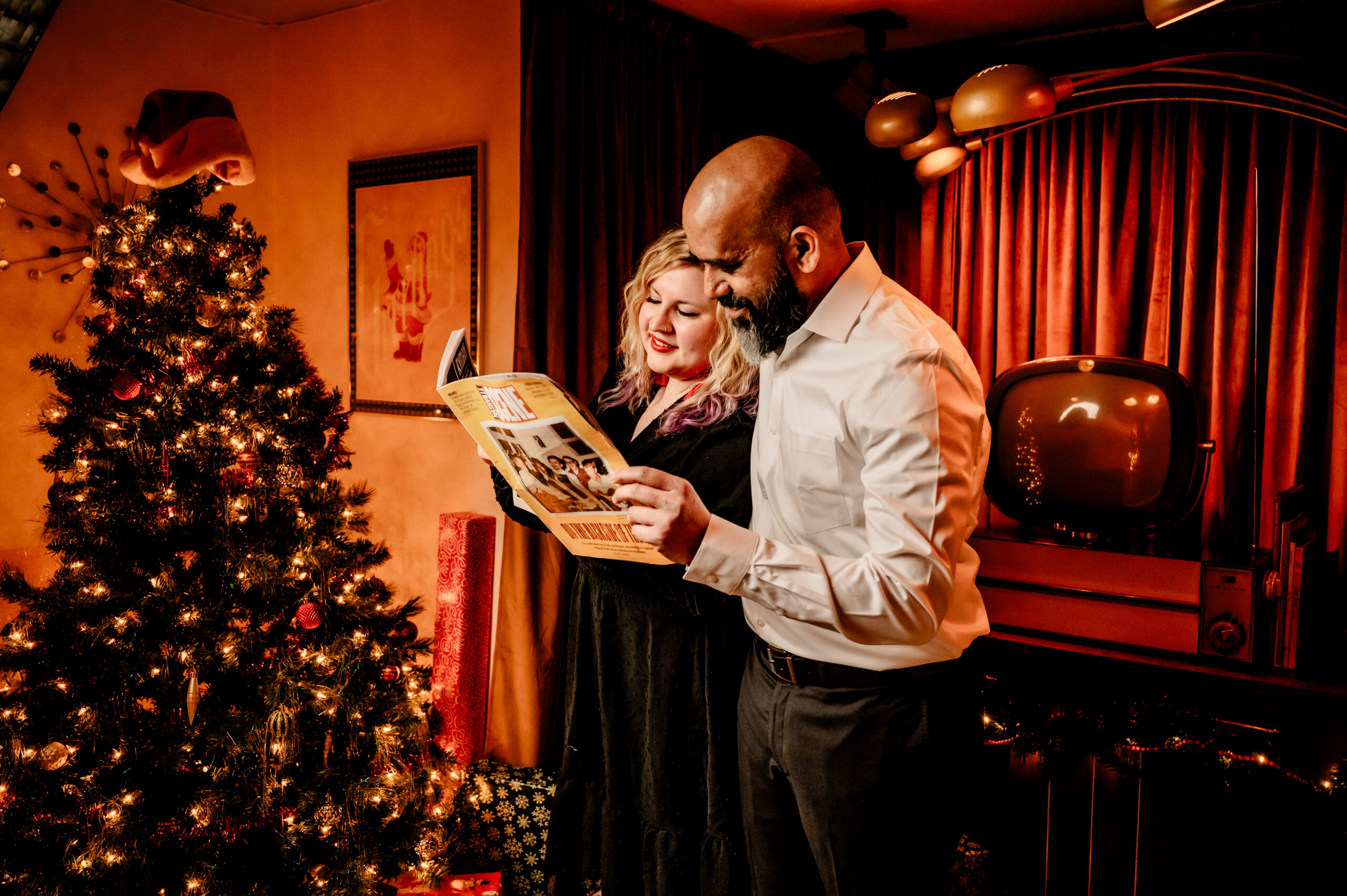 A woman and a man stand beside a decorated Christmas tree, smiling while reading a magazine together in a warmly lit, retro-styled living room with a vintage TV and festive decor—a perfect Vintage Christmas couples photography moment.