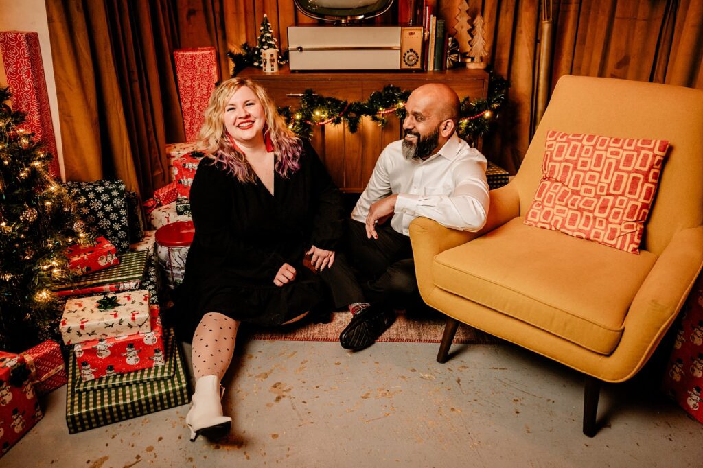 A woman and a man sit smiling on the floor by a yellow chair, surrounded by wrapped gifts and holiday decorations, with a Christmas tree and garland in the background—a perfect scene for holiday photography.
