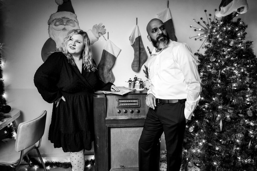 A woman in a dark dress and a man in a white shirt pose beside a vintage radio, surrounded by stockings and a glowing tree. This Vintage Christmas Holiday Couples Portrait exudes festive charm in classic black and white.